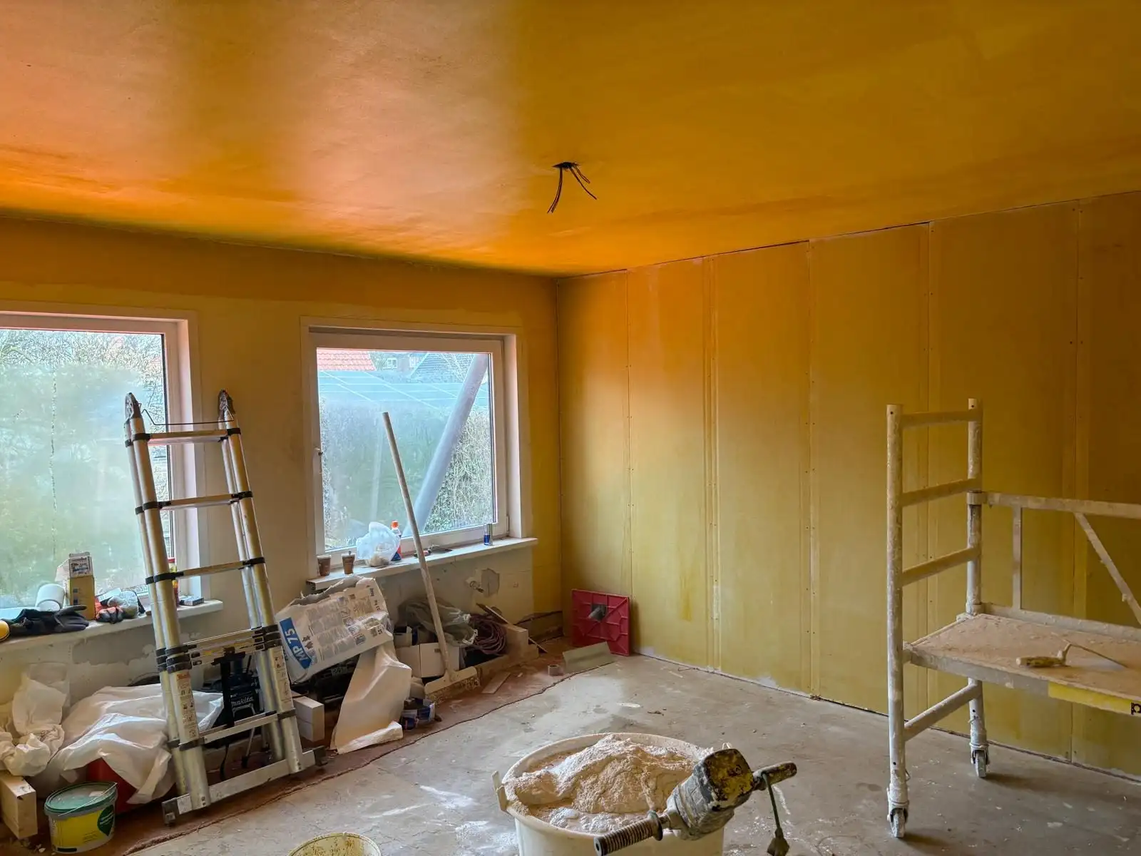 Kamer met gele voorstrijk op wanden en plafond, ladder, rolsteiger en mortelkuip.